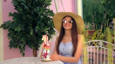 Kafedeki şık kadın geniş kenarlı şapka, güneş gözlüğü ve mavi bluz giyiyor. Renkli çok katmanlı lezzetli tatlı ya da masada milkshake.