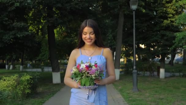 彼女の手に花の花束を持つ素敵な女性は夏に屋外の公園を歩いています — ストック動画