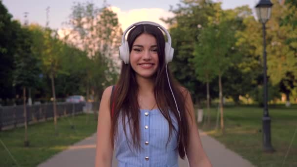 魅惑的な女性は夏に公園で屋外を歩いていて ヘッドフォンで音楽を聴いています スタイリッシュな女性は青いルーマースイートを着ています — ストック動画