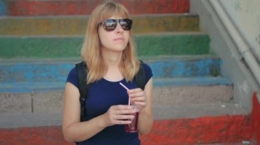 Güneş gözlüklü güzel sarışın kadın sokakta taze nar suyunun tadını çıkarıyor. Renkli merdiven arkaplanı. 