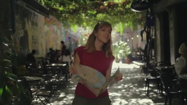 素敵な旅行者の女性は彼女の手に都市マップを保持しながら誰かに手を振っています 街中を旅する — ストック動画