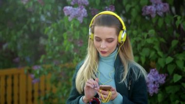 Uzun saçlı, siyah zümrüt yeşili ceketli zayıf bir kız sokakta sarı kulaklıklarla müzik dinliyor.