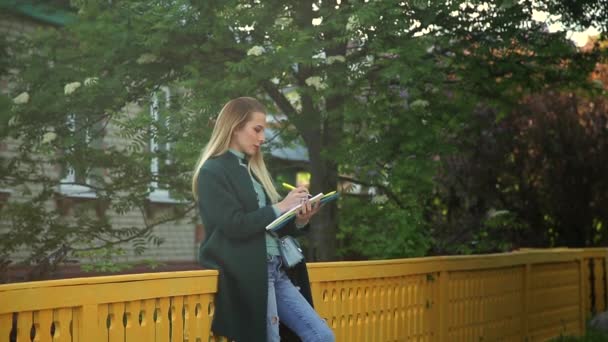 暗いエメラルドグリーンのコートを着た学生の女の子が通りのノートに書いています — ストック動画