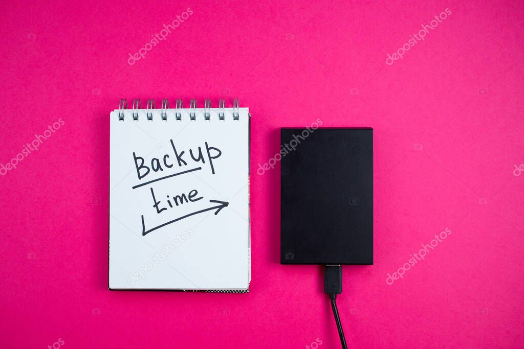 Black hard drive with a Notepad Backup text time is lying on a pink background. View from the top. Backup concept.