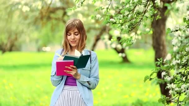 長いブロンドの髪の青いシャツに身を包んだヨーロッパの女の子の学生は ライラックのTシャツは公園の緑のノートに書き込みます 背景に白い花木 — ストック動画