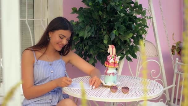 素敵なヨーロッパのブルネットの女性は 屋外カフェで時間を過ごしている間 彼女のノートに書いています テーブルの上にはカラフルなミルクシェイクとサングラスが — ストック動画