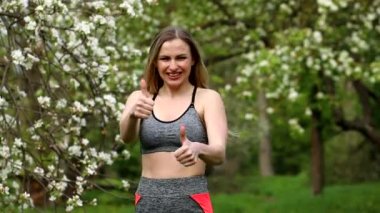 Gri eşofman giyen kız spor eğitmeni tamam diyor ve başparmağını yukarı kaldırıyor. Arka planda beyaz çiçek açan ağaçlar.