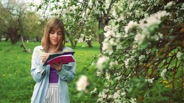 青いシャツに身を包んだ金髪の白人女子大生 ライラックブラウスが緑のノートに書いてあります 背景に白い春の開花木 — ストック動画