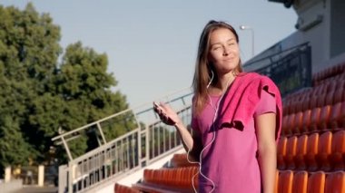 Genç çekici atletik kız stadyumda müzik dinliyor. O, pembe bir tişört giydi. Bahar güneşi yüzünü aydınlatıyor.