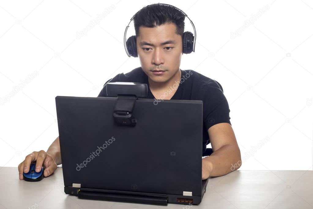 Asian male wearing headphones playing a video game and streaming online with a webcam on a laptop pc.  The image depicts entertainment industry and electronics competition or e-sports. 