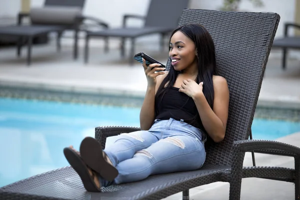 Black female on a speaker phone call in a hotel resort.  She is working while on vacation or dictating reminders on a voice assistant on her cellphone. The woman is sitting on a chair by the hotel pool.