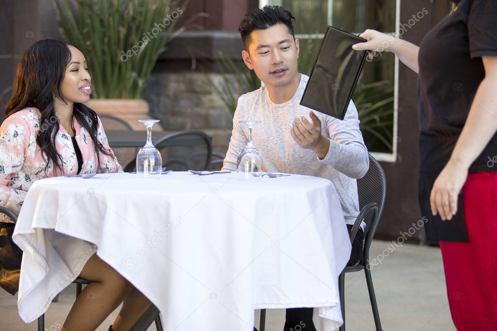 Interracial couple on a date ordering from a waiter in an outdoor restaurant.  The image depicts service industry and romantic date.