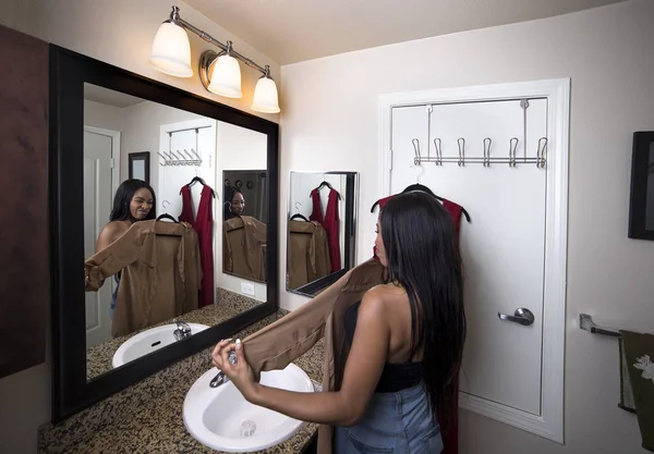Black female deciding between dresses and thinking about choosing a stylish fashion to wear.  She is getting ready to go out or trying on clothing from shopping.  The woman is looking at a mirror in the bathroom.