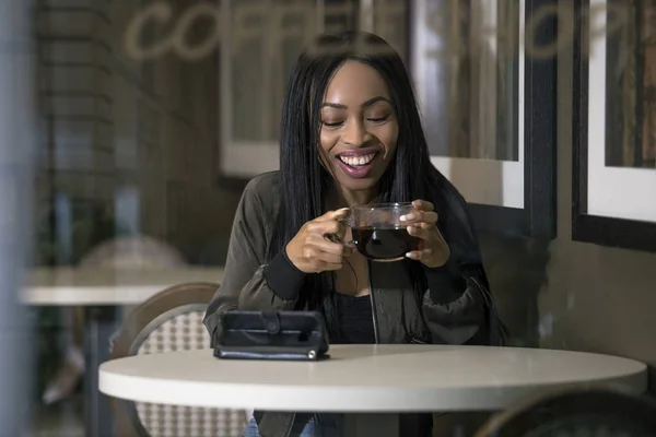 Wifi 経由でコーヒー ショップで携帯電話の黒の女性見てストリーミング ビデオを表示するウィンドウ インターネット 彼女が座っていると 一杯のコーヒーを保持しています — ストック写真