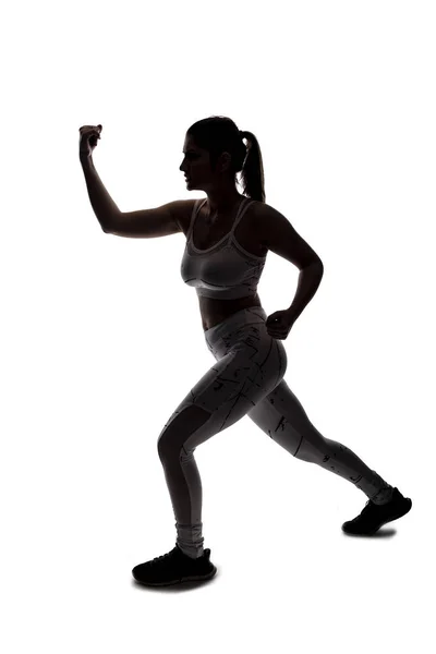 Fit young woman in a fighting stance wearing athletic sports wear and exercising by punching or practicing self defense.  She is backlit as a silhouette on a white background