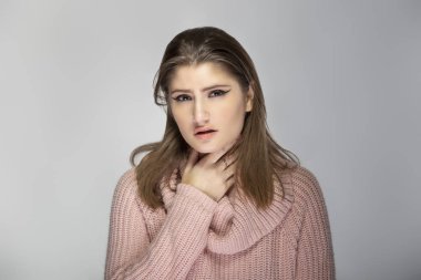 Close up portrait of a young Caucasian woman wearing a pink sweater on a grey background.  The model looks sick with a sore throat clipart