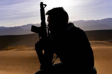 Çölün gölgesinde elinde tüfekle dinlenen bir erkek askerin silueti. Özel askeri, milis ya da özel kuvvetleri gösteriyor. Yorgun ve evini özlemiş görünüyor.