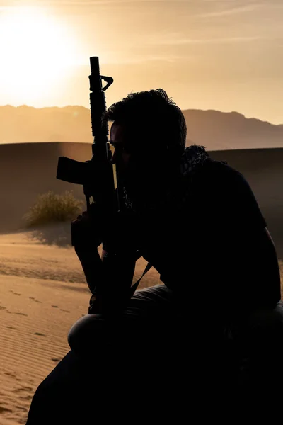 Silhouette of a male soldier resting in the shade on a desert and holding a rifle.  Depicts the private military industry, militia, or special forces.  He looks tired and homesick