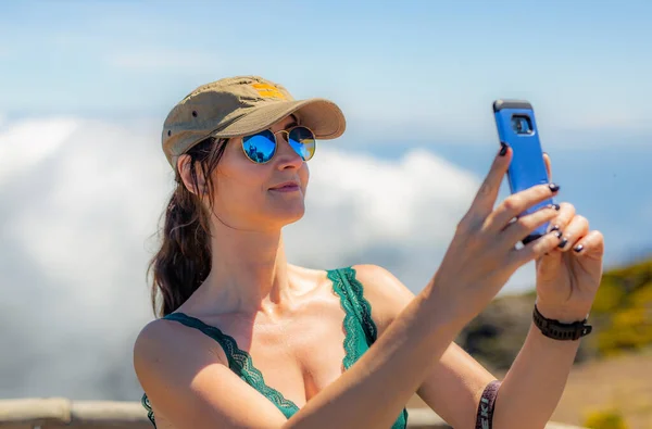 Şapkalı ve güneş gözlüklü bir kadın bulutların arkasında selfie çekiyor.