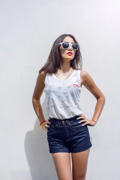 Chica morena en verano en el parque con descanso en pantalones cortos y una blusa blanca en el estilo de vida de moda de fondo, aspecto suave. Maquillaje sensual, piel bronceada lápiz labial rojo brillante. En gafas de sol . — Foto de Stock