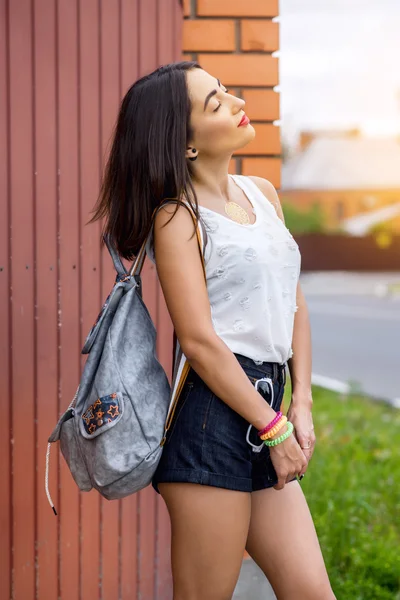 Chica morena en verano en el parque con una mochila descansando en pantalones cortos y blusa blanca, estilo de vida de moda, mirada suave. Maquillaje sensual, piel bronceada brillante . — Foto de Stock