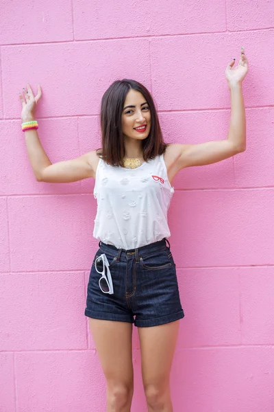 Schöne brünette Mädchen in weißer Bluse und Jeans Shorts Spaß auf dem Hintergrund einer rosa Wand, lächelnd, Urlaubskonzept, Lifestyle, zeigt seine Hände nach oben. — Stockfoto