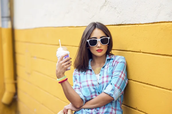 Femme dans la ville sur le fond d'un mur jaune, tenant milkshake, jus frais dans des lunettes, portant chemise avec les bracelets son bras, makiyah lèvres rouges sensuelles, style de vie concept de loisirs — Photo