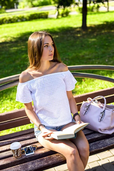 Hermosa chica morena sentada en un banco en el soleado parque de verano, leyendo libro, disfrutar de sus vacaciones, concepto de estudiante, después de la escuela en el recreo, descansando con la taza de café o té teléfono inteligente —  Fotos de Stock