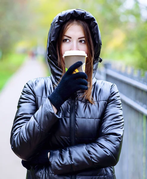 Ragazza in giacca con cappuccio, in possesso di un caffè o tè, giovane gode la vita all'aria aperta e sport, primavera autunno, stile di vita, nero, il concetto di colazione, guanti — Foto Stock