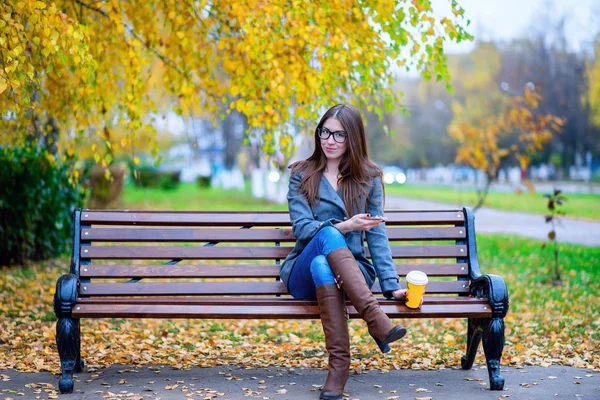 Fille en veste assis sur le banc avec des lunettes, tenant un café ou un thé, jeune en plein air, printemps automne, style de vie, le concept de la ville, style de vie, sourire, heureux. le téléphone en main . — Photo