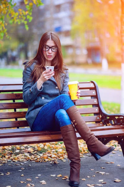 Meisje leest sms, herschreven smartphone buitenshuis in de lente of herfst, op de Bank, met koffie-thee, Ontbijt, concept, brillen, urban fashion, lifestyle, brunette. — Stockfoto