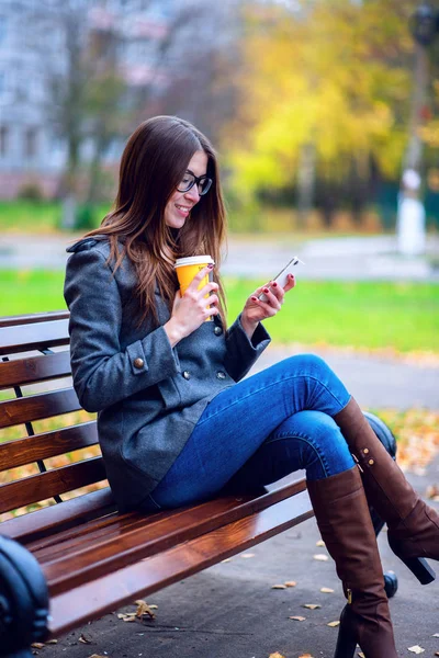 Girl membaca sms, ditulis ulang smartphone luar ruangan di musim semi atau musim gugur, di bangku, dengan teh kopi, sarapan, konsep, kacamata, mode perkotaan, gaya hidup, brunette. Selamat tersenyum . — Stok Foto