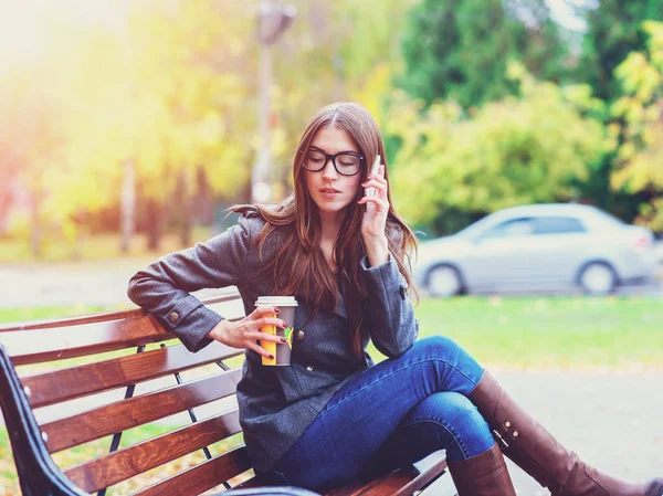 Chica con chaqueta hablando por teléfono, sentado en un banco, sosteniendo café o té, al aire libre, otoño de primavera, estilo de vida, el concepto de la ciudad, estilo de vida, escuchando la conversación . — Foto de Stock