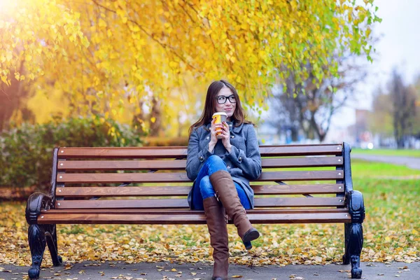 Schöne junge Frau, die auf einer Bank sitzt und Kaffee oder heißen Tee im Frühjahrsmantel trinkt und im Park im Freien genießt, Brille, urbanes Leben, das Konzept des Frühstücks in der Natur — Stockfoto