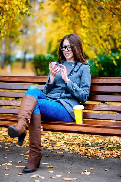 Ragazza in giacca seduta sulla panchina con gli occhiali, tenendo un caffè o un tè, giovani all'aperto, autunno primaverile, stile di vita, il concetto di città, stile di vita, sorriso, felice. il telefono in mano . — Foto Stock