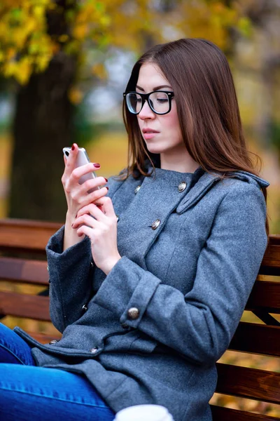 Meisje leest sms, herschreven smartphone buitenshuis in de lente of herfst, op de Bank, met koffie-thee, Ontbijt, concept, brillen, urban fashion, lifestyle, brunette. — Stockfoto