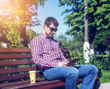 A man on a bench in a city park, read a message on the tablet, watching a movie or TV show, next to a glass of coffee or tea, holiday lunch outdoors. clipart