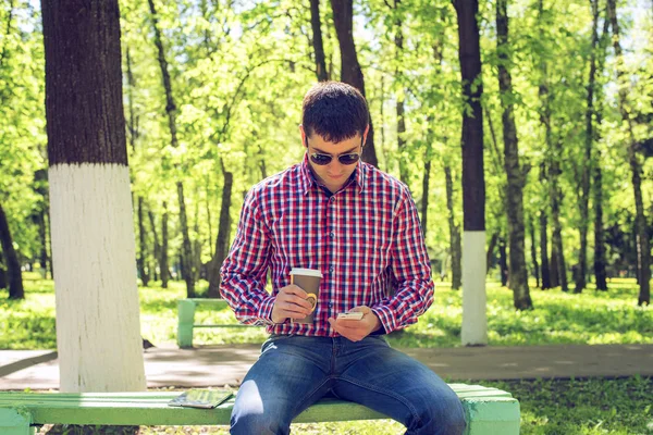 Der Mann auf der Bank, mit Kaffee oder Tee, liest eine Nachricht am Telefon, im Sommer in Brille und Hemd im Wald im Freien ruht Geschäftsmann. — Stockfoto