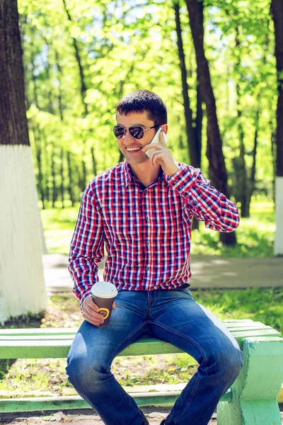 Der Mann auf der Bank, mit Kaffee oder Tee, telefonierend, im Sommer in Brille und Hemd im Wald im Freien, glücklich lächelnd Geschäftsmann. — Stockfoto