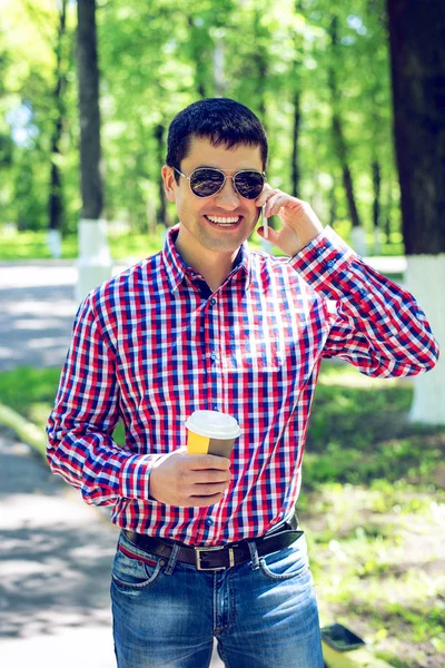 Mann mit Kaffee oder Tee, telefonierend, im Sommer in Brille und Hemd im Wald im Freien, glücklich lächelnd Geschäftsmann, der Emotionen spricht. — Stockfoto