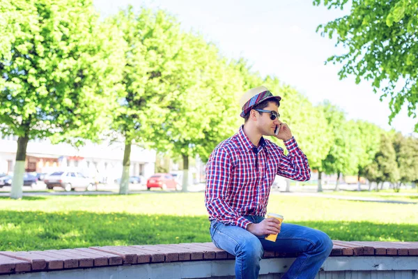 Der Mann sitzt, ein Smartphone in der Hand, telefoniert, im Sommer in Brille und Hemd mit Hut auf einer Bank im Wald und ruht sich aus. — Stockfoto