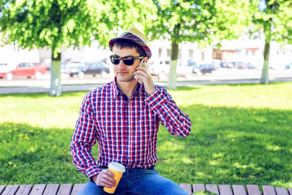 Der Mann sitzt, hält einen Kaffee oder Tee in der Hand, telefoniert, im Sommer in Brille und Hemd mit Hut im Wald, im Freien ruht ein Geschäftsmann und telefoniert mit dem Smartphone.. — Stockfoto