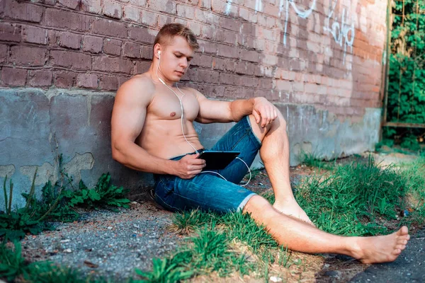 Um atleta masculino moderno em shorts está sentado na rua com um tablet e fones de ouvido, assistindo a um filme em redes sociais desfrutando de recreação ao ar livre, no verão em uma parede de tijolo, conceito horizontal — Fotografia de Stock