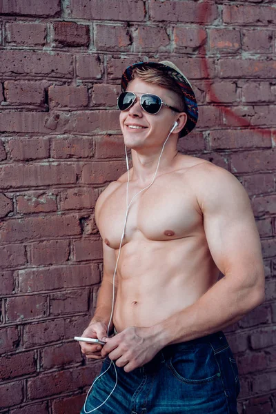 A happy male athlete, smiling in fresh air wearing a hat and glasses, with a smartphone and headphones listening to music, a fun outdoor recreation, an inflated bodybuilder in summer at wall, a — Stock Photo, Image