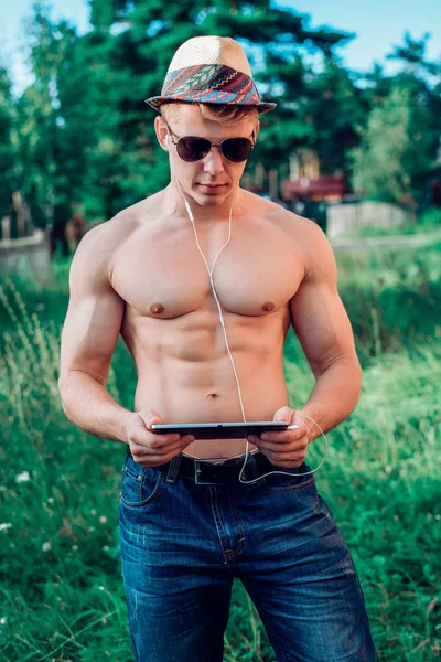 Un hombre moderno guapo, un atleta chico de culturistas está de pie en el parque en la naturaleza un montón de vegetación, en jeans con una tableta auriculares, viendo vídeo en las redes sociales. En gafas un musculoso — Foto de Stock