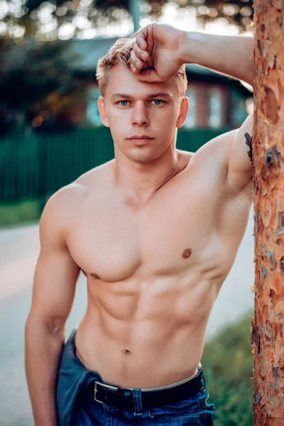 Un hombre en verano por árbol, primer plano de la naturaleza, en jeans un deportista trabajador, concepto de un hombre moderno vertical, descansando. La piel bronceada es una prensa muscular. Una mirada de retrato segura. Un pecho inflado — Foto de Stock