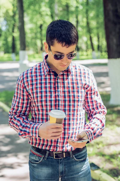 Um homem de camisa e jeans e óculos de sol, segurando um celular café ou chá, ler as mensagens de conversa em seu telefone, o conceito de verão, um homem de negócios em férias. Estilo de vida da cidade. Sobre o — Fotografia de Stock