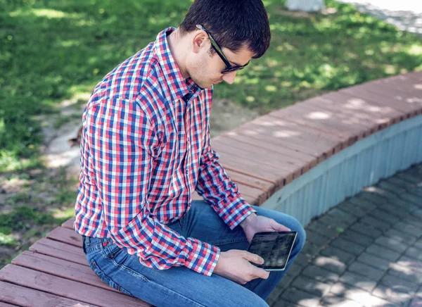 man in  shirt and jeans  sunglasses, video looks on the tablet corresponds to the social networks, in the summer  the bench, the concept of  businessman  vacation. City lifestyle.  the street in th