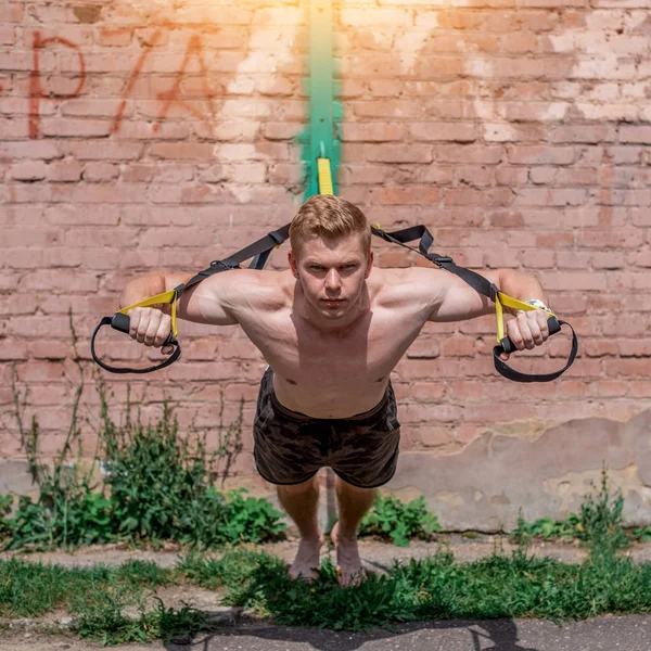 A male athlete is engaged in open air loops, trains chest, pushing away from brick wall. In summer, in shorts and a bare torso. Healthy lifestyle of young people. The concept of sport motivation