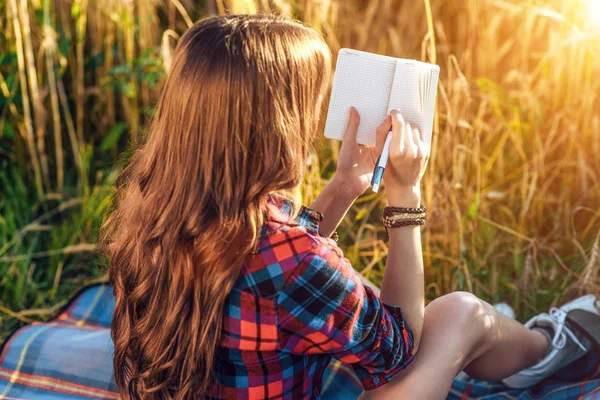 Meisje zit in een veld shirt, tarwe ontspannen aard, mooi haar. Schrijft ideeën in een notitieblok. Een student na school. Concept nota 's. — Stockfoto